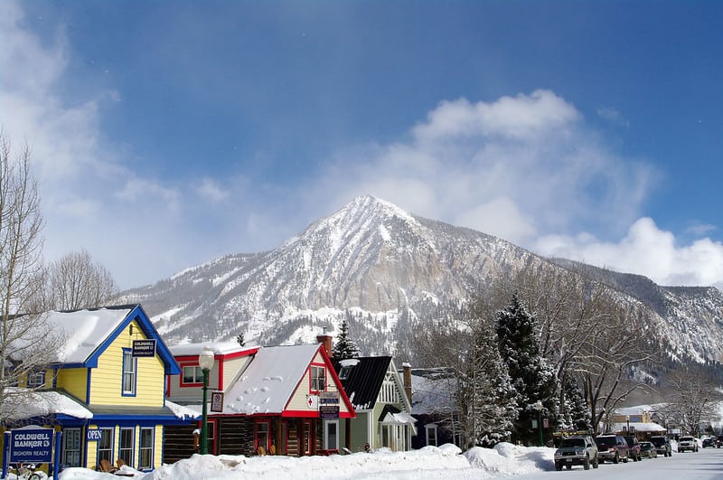 A row of small houses stand in deep snow, with a snowy peak of the Rocky Mountains in the background.