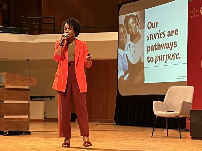 Photo by Becky McCray. Slide by Kia Weatherspoon A woman speaking on stage. She has dark skin and dark hair in a natural style. The slide on screen shows her as a child with her brother, and the text says, "Our stories are pathways to purpose."