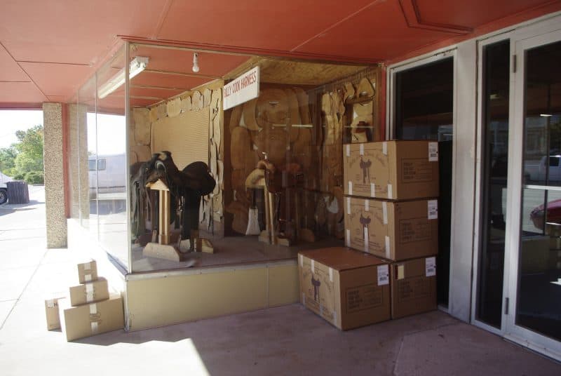 A repurposed downtown retail building is used for storage and warehousing by a saddle manufacturer. A window display shows saddles and templates used to make them. A sign says "Billy Cook Harness." Several boxes of saddles and accessories are stacked up outside ready for pickup by the shipping company.