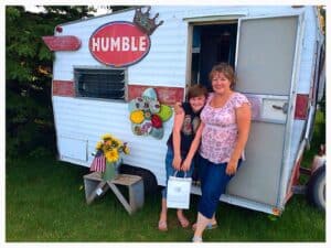 A tiny travel trailer being used as a mobile retail store, two customers are smiling just outside with their purchase.